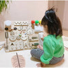 A child is interacting with a wooden Montessori busy board set featuring gears, buttons, and puzzle elements.
