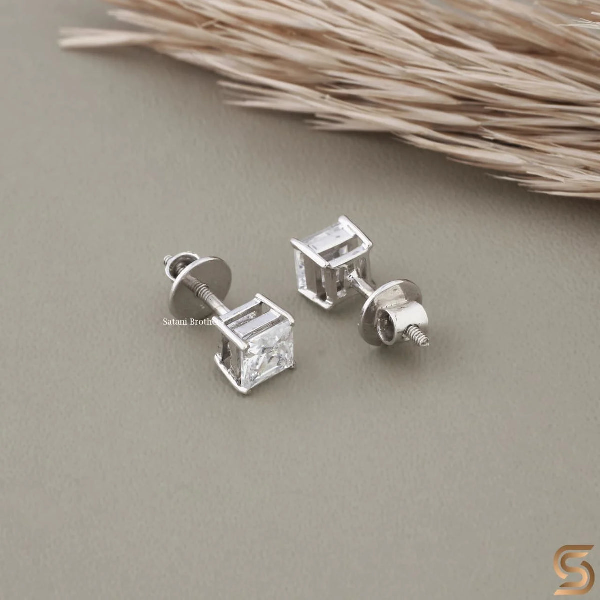 A pair of princess cut moissanite stud earrings in sterling silver resting on a neutral gray surface.