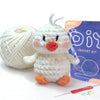 A white crocheted duck kit with orange feet and a ball of white yarn next to a purple bag.