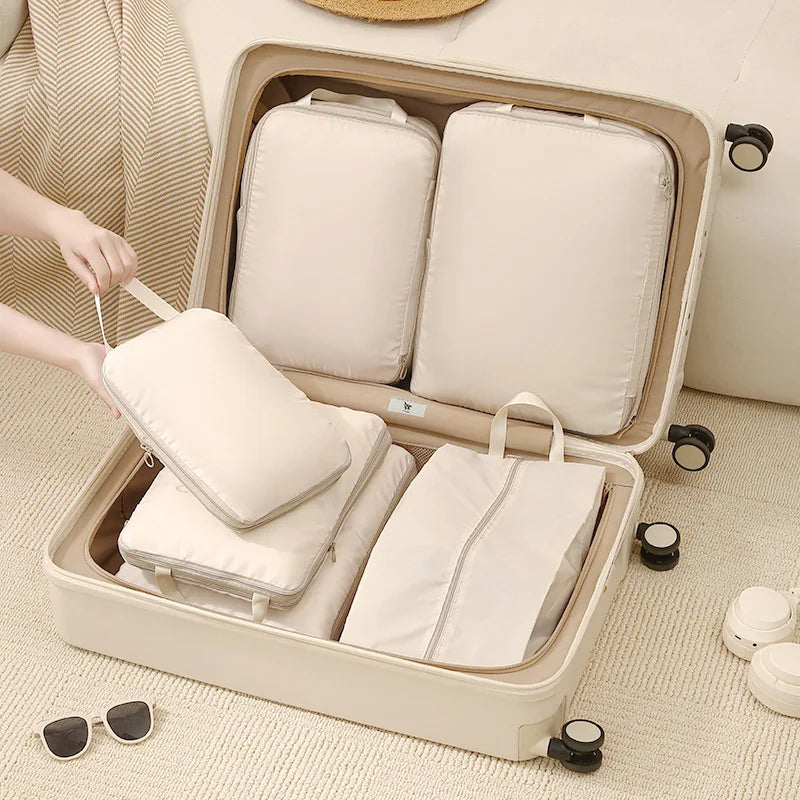 A beige packing cubes set is displayed inside a light-colored storage box with various fabric items and sunglasses nearby.