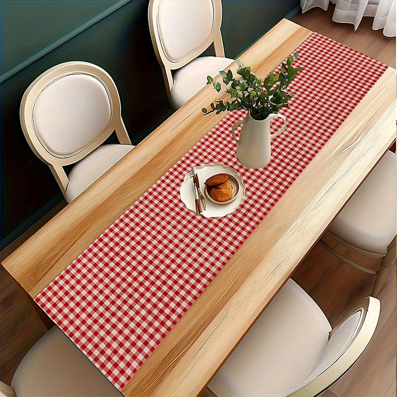A light wood table is set with a red and white gingham table runner, plates, and a vase of greenery.