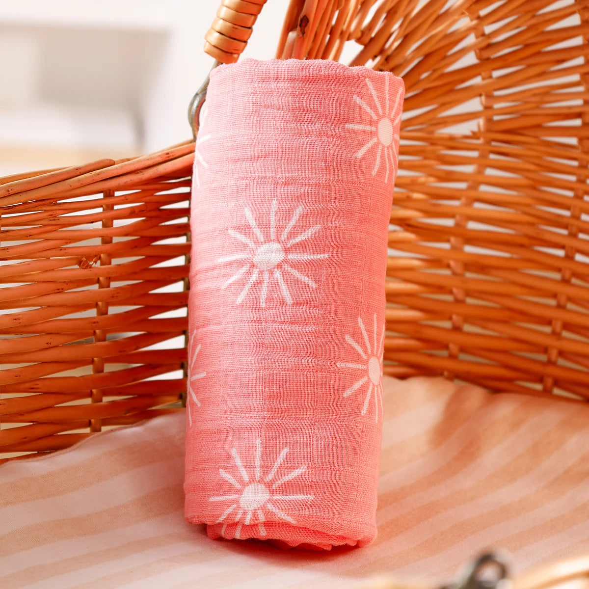 A rolled pink muslin baby swaddle blanket with white sunburst patterns is displayed in a wicker basket.