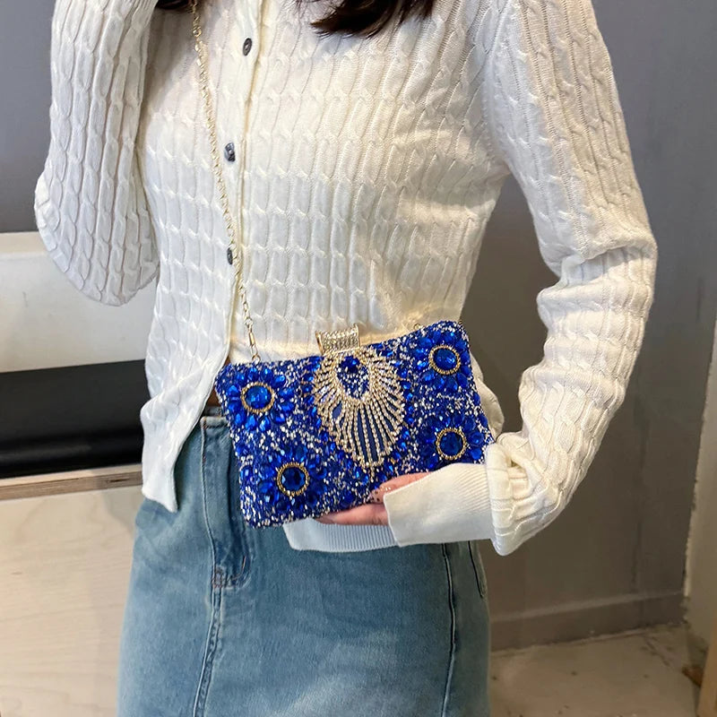 A white knitted sweater sleeve shows a blue rhinestone evening clutch with a gold chain strap and blue sequins.