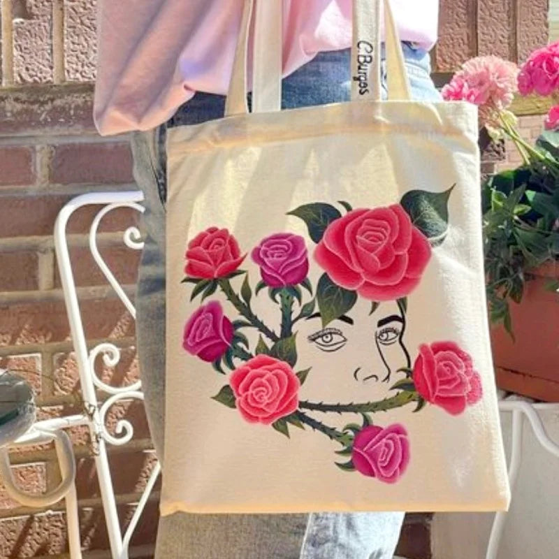 A beige canvas shopping tote bag featuring a detailed floral design of pink and red roses.
