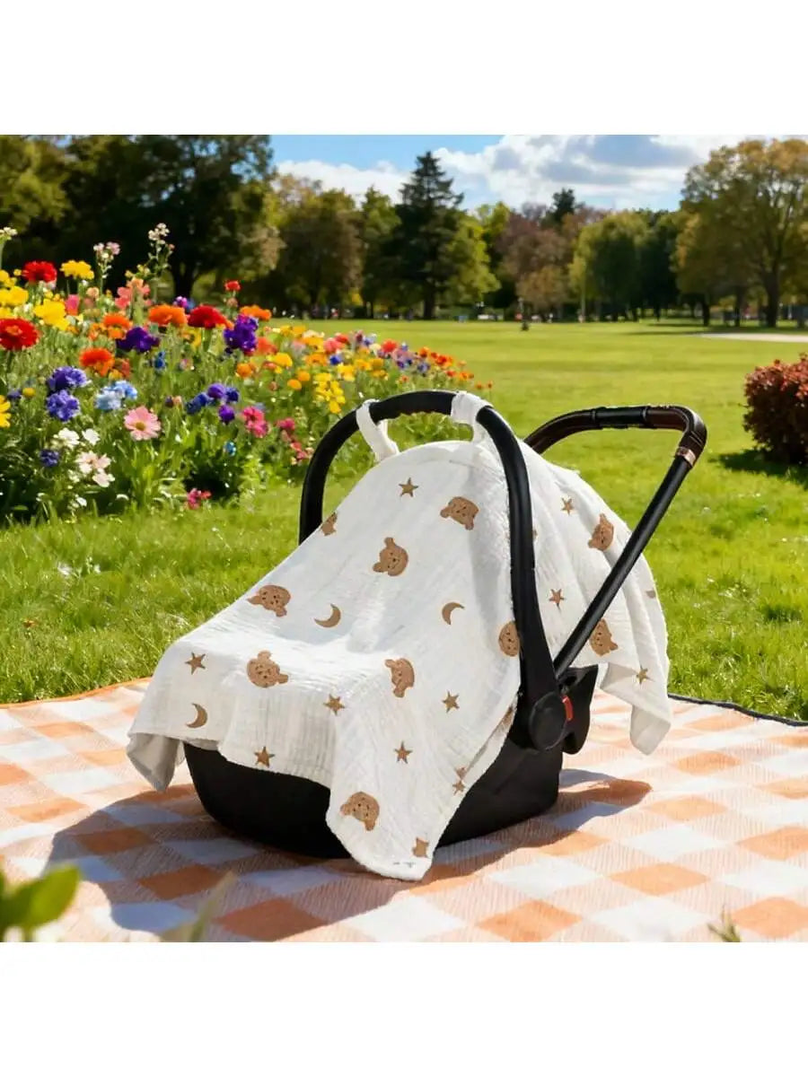 A white stroller cover with gold stars and moons is displayed on an orange checkered mat in a grassy park setting.