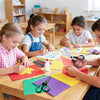 A boy and a girl are using multipurpose stainless steel scissors to cut colored paper at a wooden table.