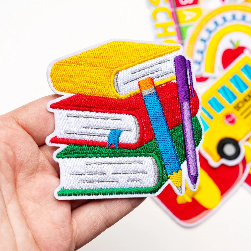 A hand holds a stack of ten colorful back to school iron-on patches featuring books, pencils, and school supplies.