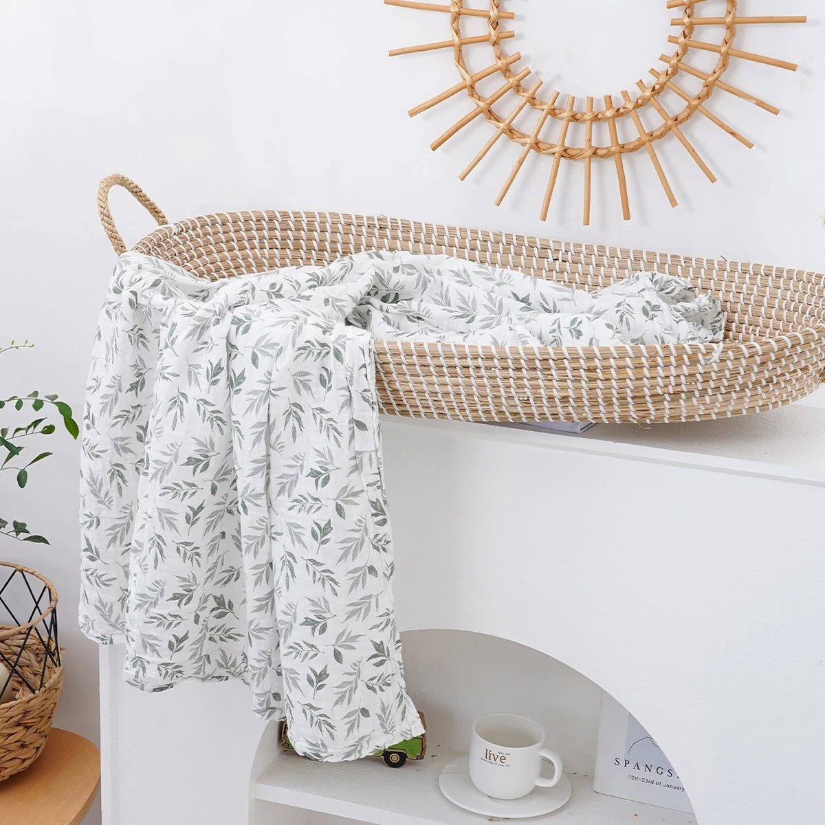 A white and green patterned muslin baby swaddle blanket draped over a woven basket on a white shelf.