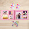 A pink scrapbook album with colorful patterned paper and decorative stickers is shown on a wooden surface.