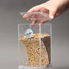A hand holds a clear plastic feeder containing bird seed, with a transparent lid above it.