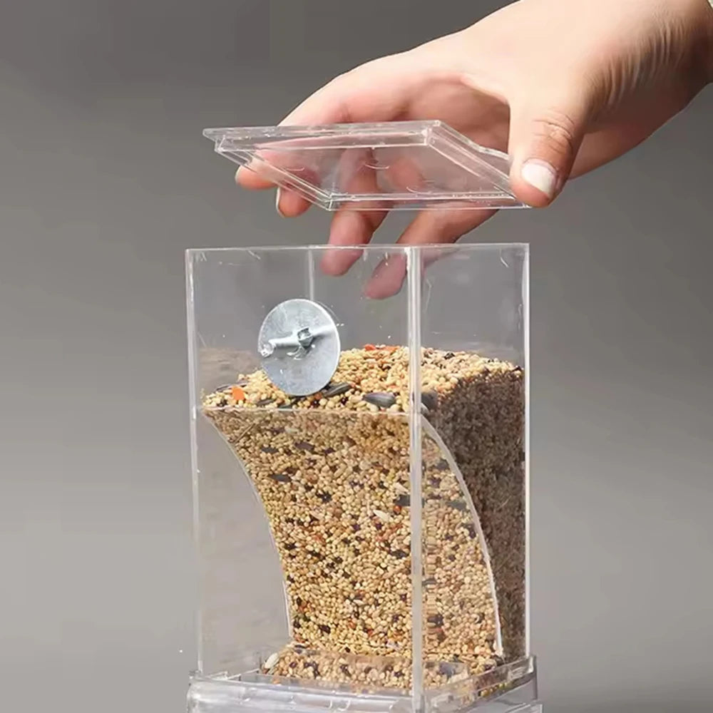 A hand holds a clear plastic feeder containing bird seed, with a transparent lid above it.