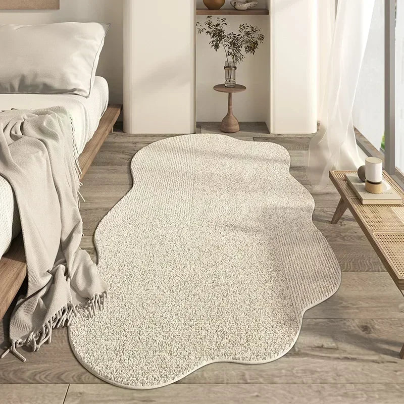 A light beige irregular area rug with a plush texture placed on a wooden floor next to a white wall and bedding.