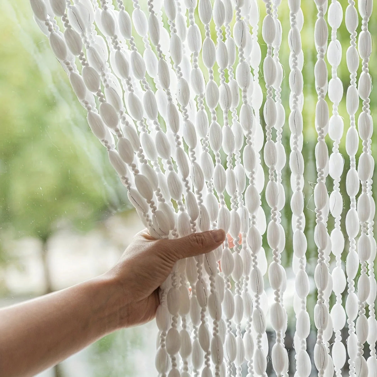 A hand touches a white bohemian tassel curtain with lanterns filtering light through the fabric.