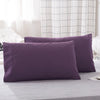 A set of two solid purple microfiber pillowcases resting on a gray bed with a white checkered curtain in the background.