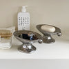 A silver ceramic soap dish with an elevated stand holds a round bar of soap in front of a white bottle.