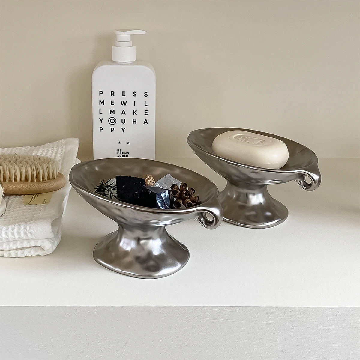A silver ceramic soap dish with an elevated stand holds a round bar of soap in front of a white bottle.
