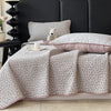 A light gray floral quilted bedspread with pink trim is draped over a dark bed, next to a white stuffed animal pillow.