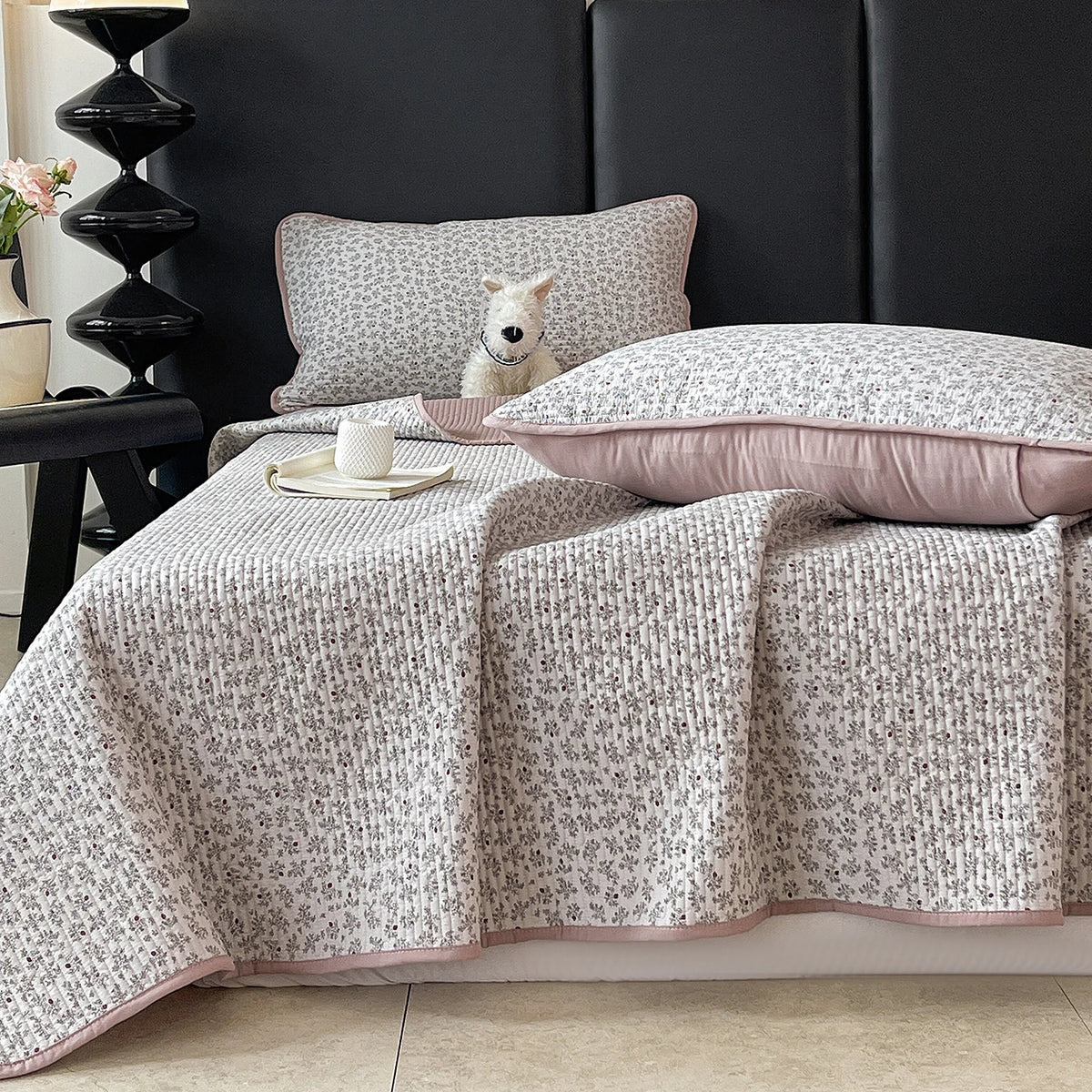 A light gray floral quilted bedspread with pink trim is draped over a dark bed, next to a white stuffed animal pillow.