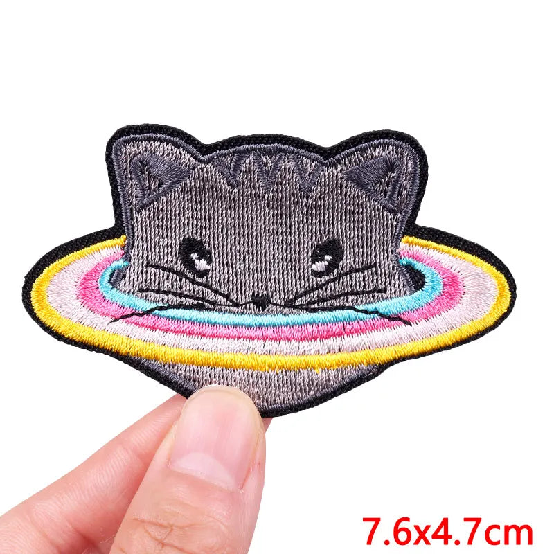 A gray cat face embroidered patch with a colorful rainbow border being held by a hand.