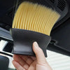 A hand holds a black car interior detailing brush set with soft yellow bristles, positioned inside a car vent.