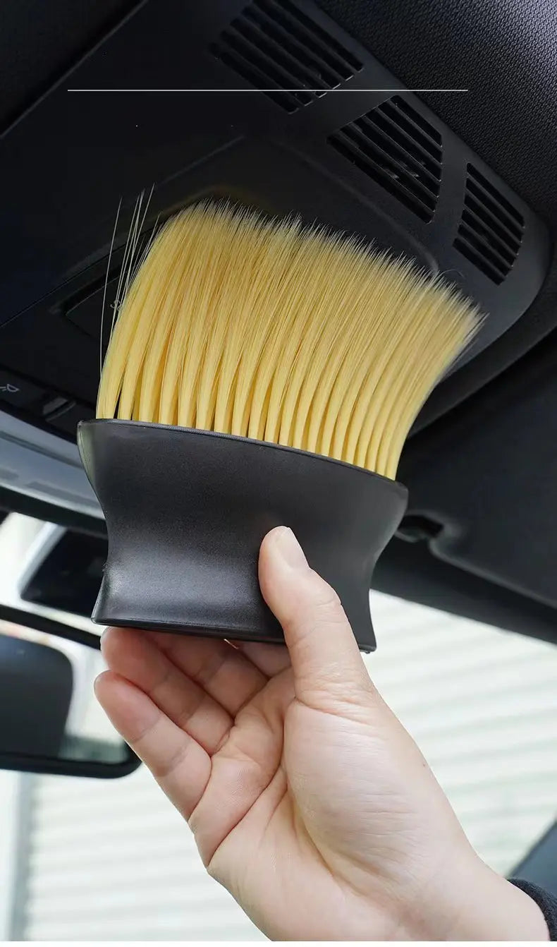 A hand holds a black car interior detailing brush set with soft yellow bristles, positioned inside a car vent.