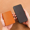A hand holds a black smartphone and an orange PU leather compact wallet against a brown background.