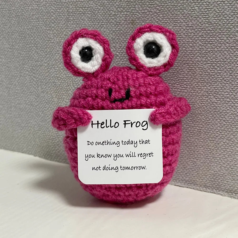 A handmade crochet frog doll with large eyes and a smiling face, knitted in pink yarn.