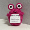 A handmade crochet frog doll with large eyes and a smiling face, knitted in pink and white yarn.