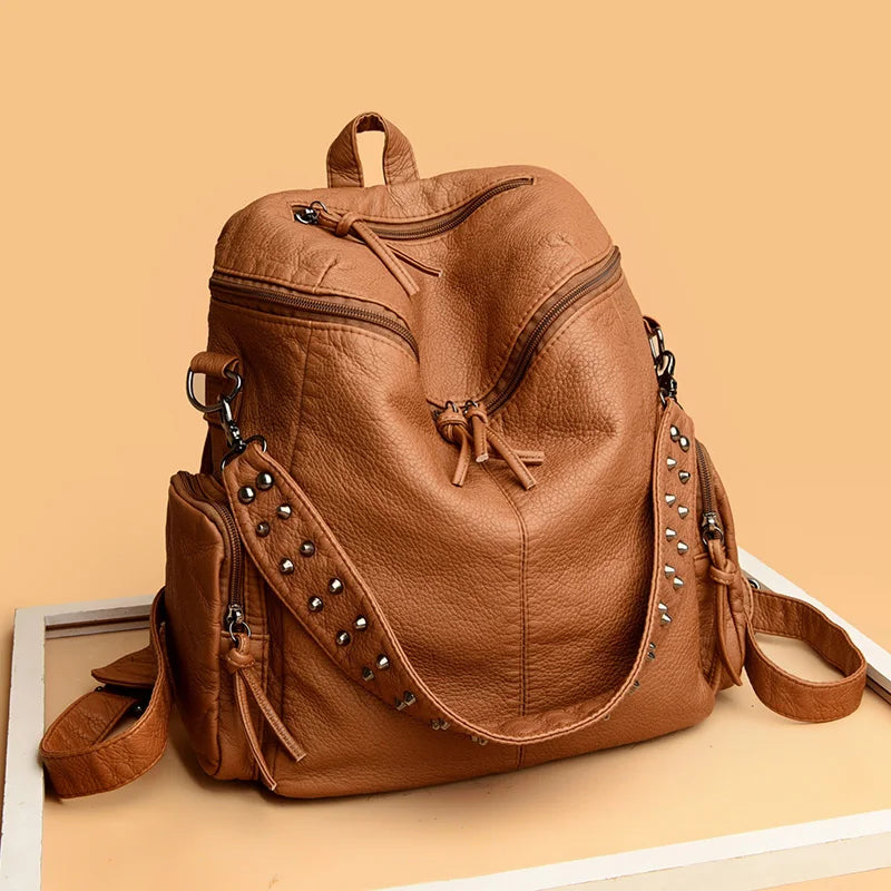 A tan leather backpack with rivets and drawstring closure is shown on a light surface.