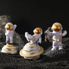 Three handcrafted astronaut figurines in white and gold resin standing on black gravel.