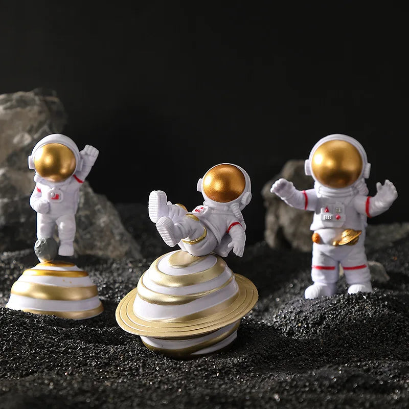 Three handcrafted astronaut figurines in white and gold resin standing on black gravel.