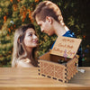 A young couple looks at a vintage hand-cranked wooden music box labeled Can't Help Falling in Love.