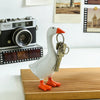 A white resin goose sculpture with orange feet holds a set of antique-style keys on a wooden surface.