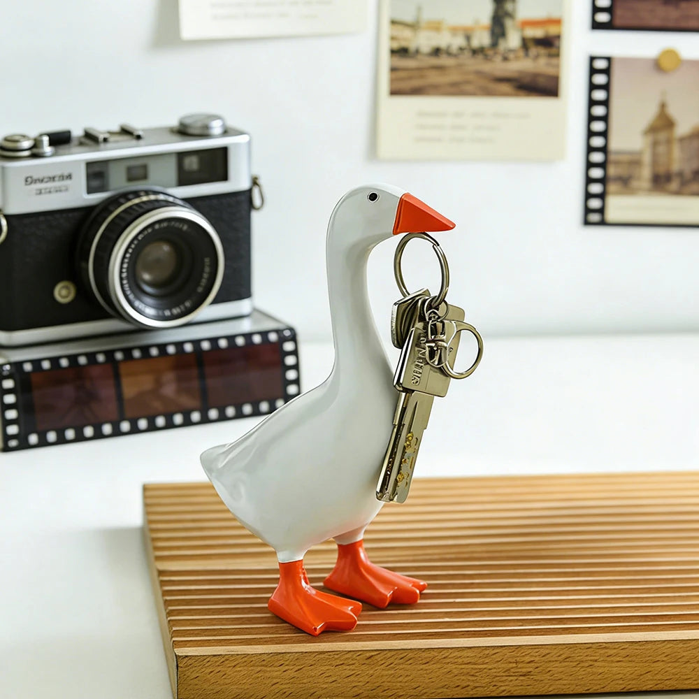 A white resin goose sculpture with orange feet holds a set of antique-style keys on a wooden surface.