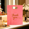 A pink gift bag with a handle and gold lettering that reads Thank you, displayed on a white surface next to perfume bottles.