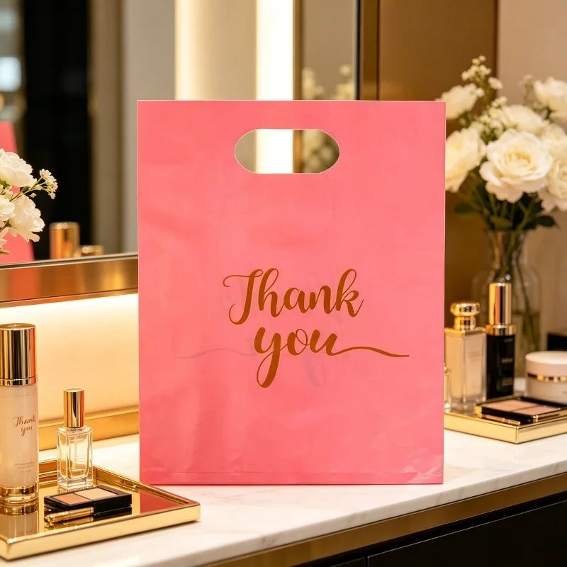 A pink gift bag with a handle and gold lettering that reads Thank you, displayed on a white surface next to perfume bottles.