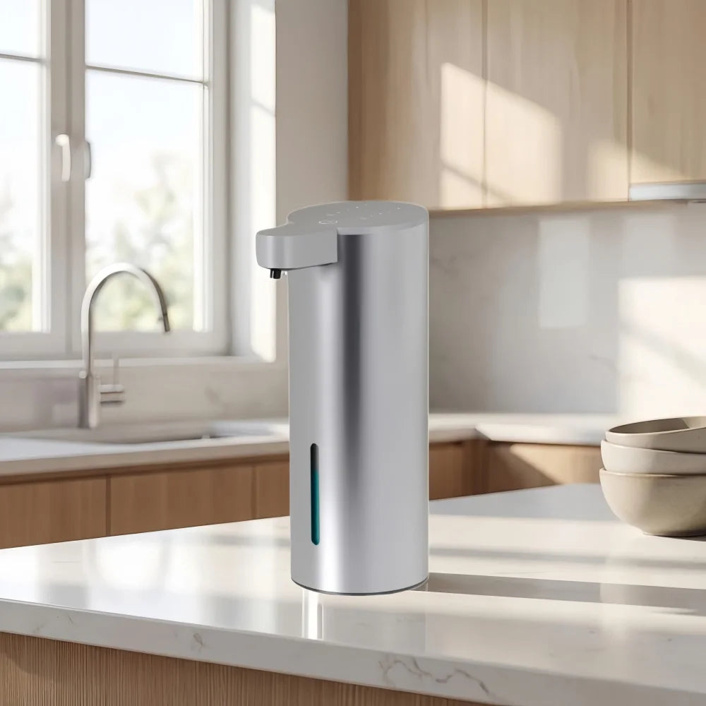 A modern stainless steel automatic foam soap dispenser sitting on a kitchen counter next to a window.