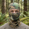 A man wearing a camouflage balaclava cap in a forest setting.