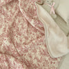 A soft pink floral muslin baby blanket with white accents and a fluffy cream-colored stuffed animal.