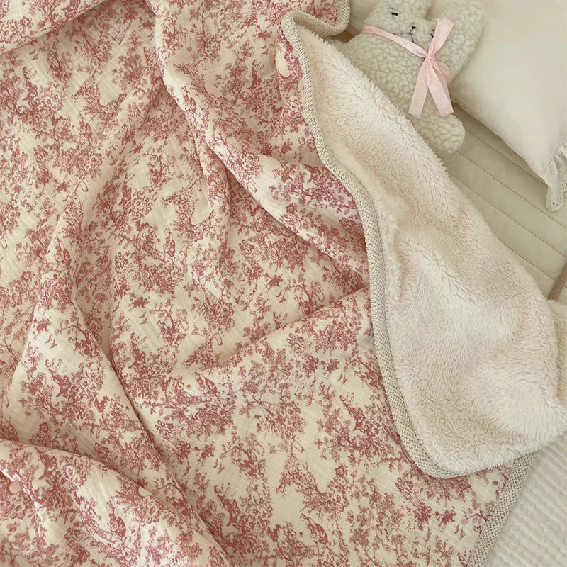 A soft pink floral muslin baby blanket with white accents and a fluffy cream-colored stuffed animal.