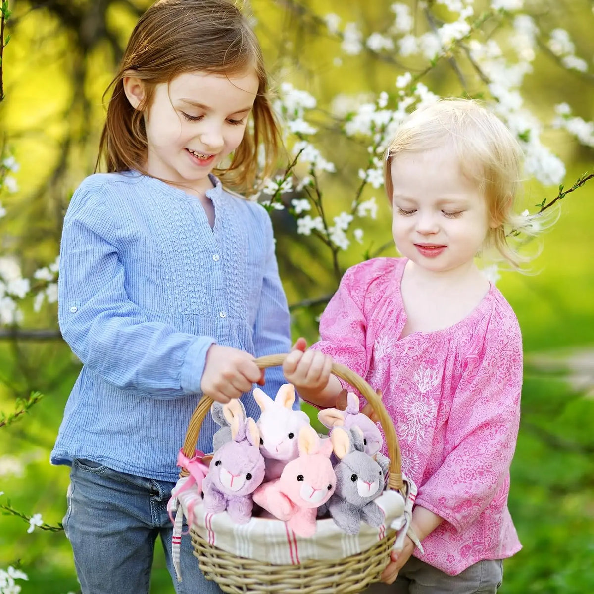 Two young girls smiling while holding a woven basket filled with pink and gray mini plush bunny toys in a sunny outdoor setti