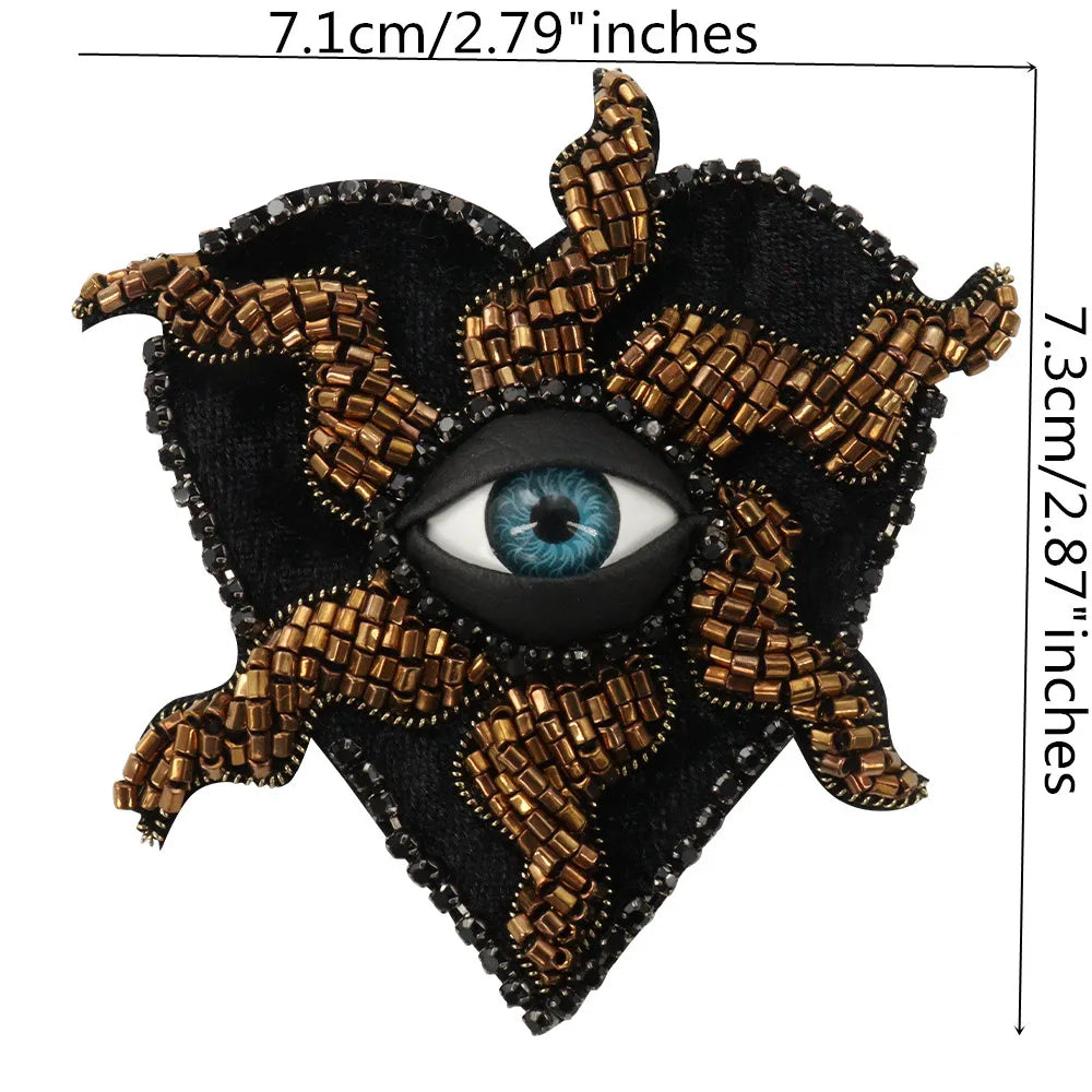 A black heart-shaped patch featuring a detailed eye design made with gold and bronze beaded rhinestones.