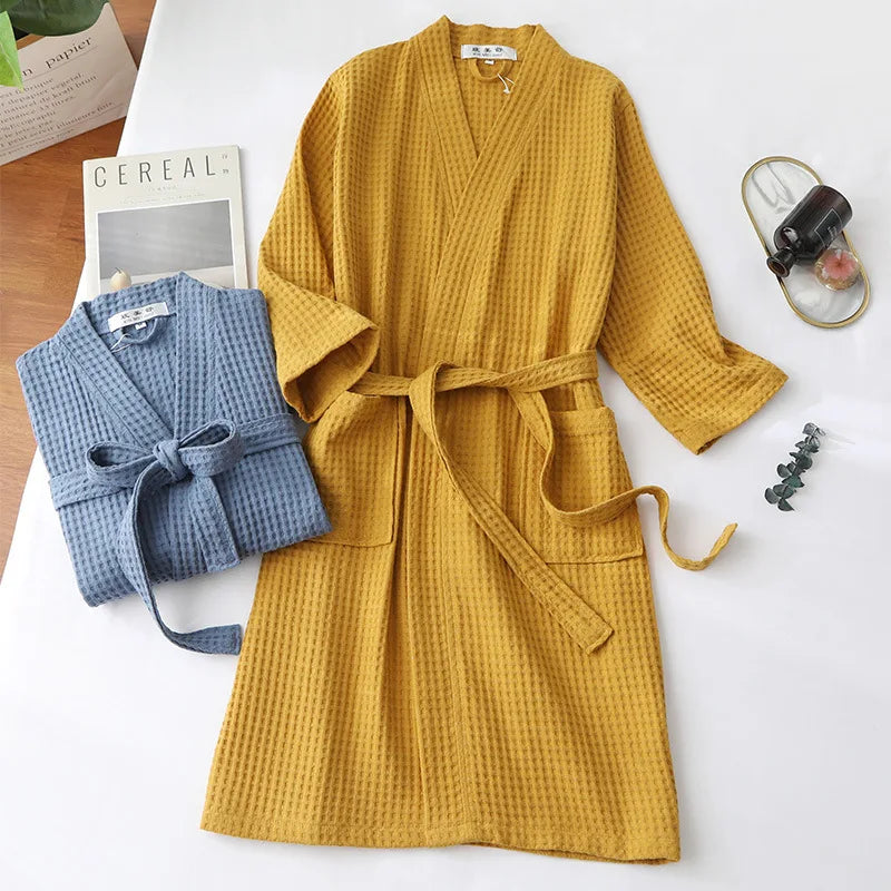 A mustard yellow lightweight cotton waffle robe hanging on a white surface next to a folded blue knitted garment.