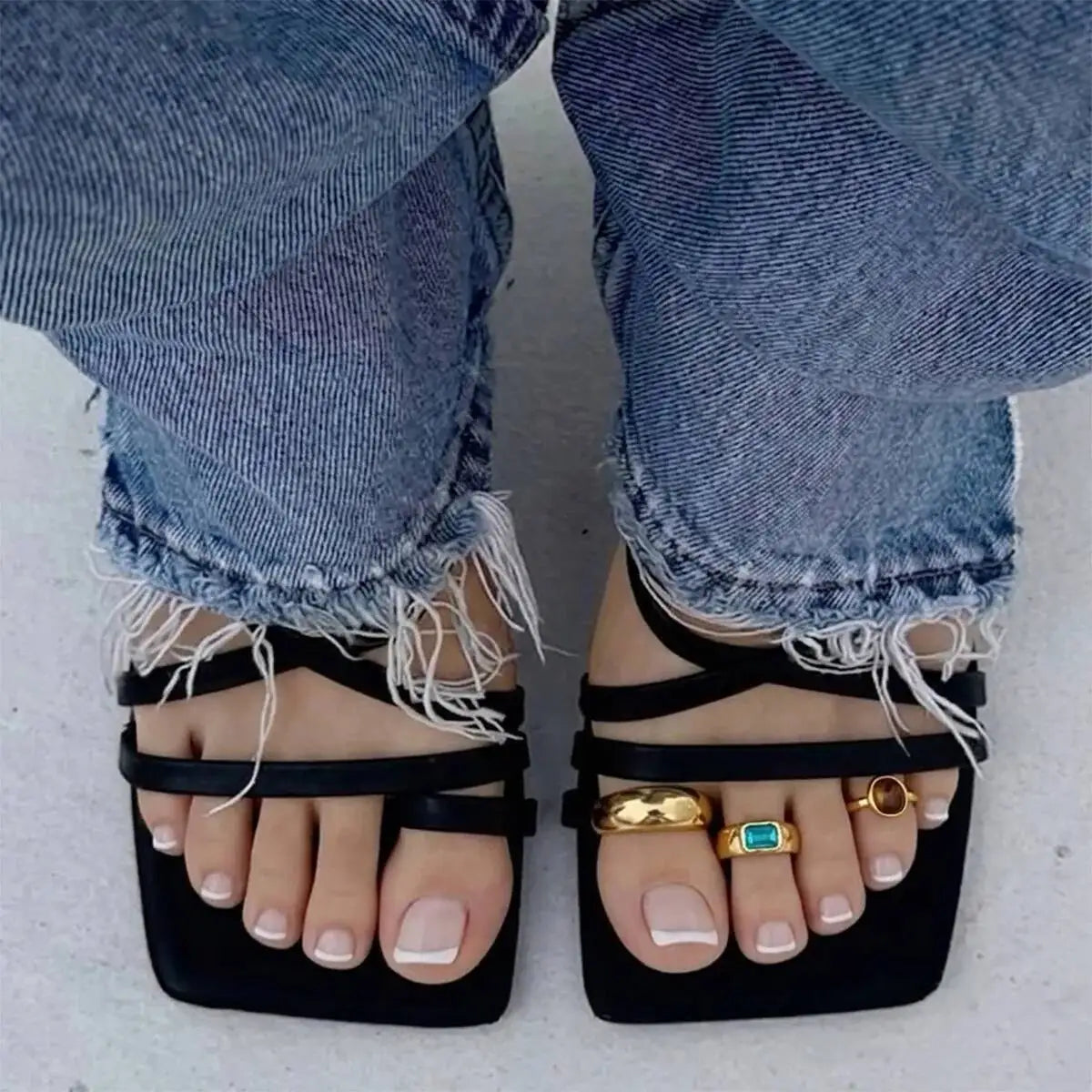 Pieds portant des sandales noires avec des anneaux à l'orteils or et turquoise, visibles à travers un jean déchiré.