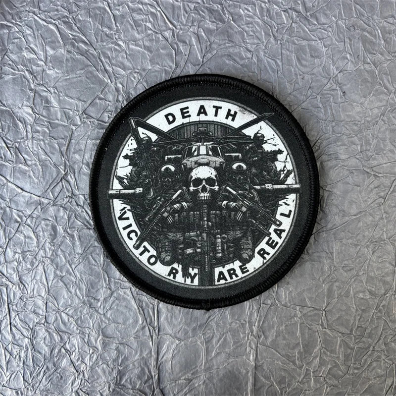 A black tactical patch featuring a skull design and the text DEATH VIC TO RY ARE REAL on a silver background.
