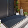 A dark blue non-slip doormat placed on a concrete walkway in front of a wooden wall with a potted plant.