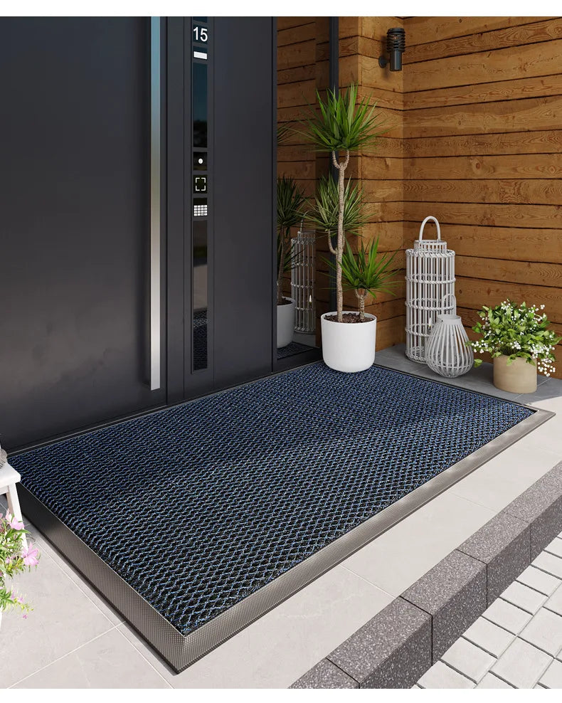 A dark blue non-slip doormat placed on a concrete walkway in front of a wooden wall with a potted plant.