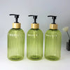 A set of three clear green glass refillable soap dispensers with black pump tops and wooden lids.