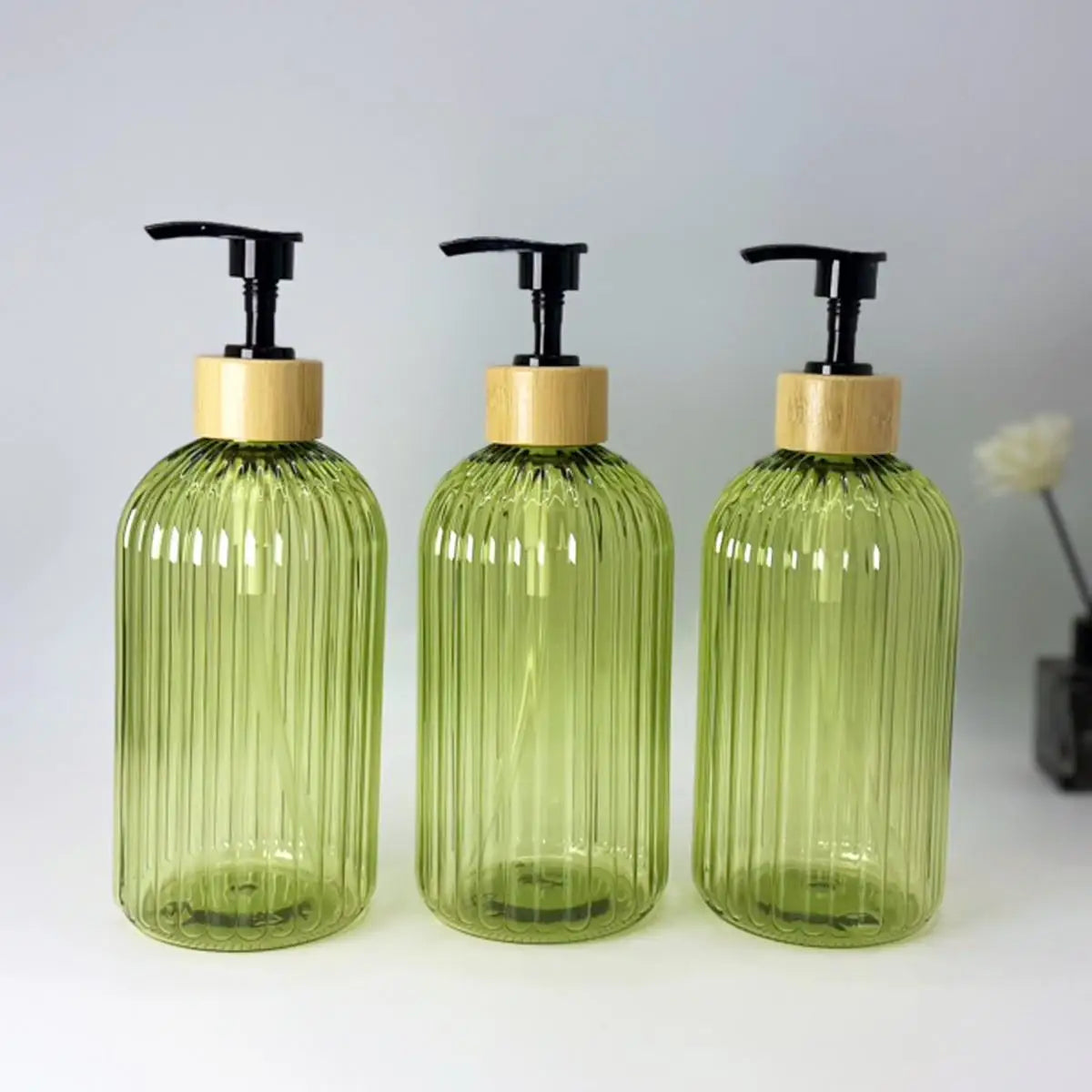 A set of three clear green glass refillable soap dispensers with black pump tops and wooden lids.