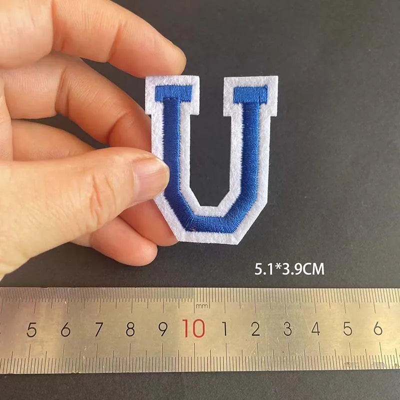 A hand holds a blue embroidered letter U patch against a ruler showing its dimensions.